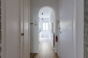 Un couloir étroit aux murs blancs et au plancher en bois, qui mène à un espace cuisine doté d’une fenêtre et d’une porte au fond.