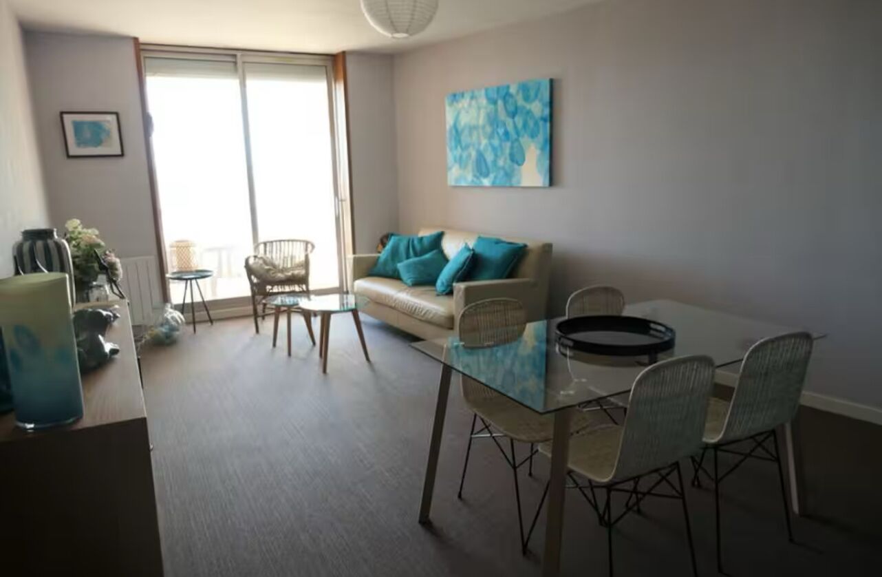 Ein modernes Wohnzimmer mit einem Glastisch, zwei Stühlen, einer Couch mit türkisfarbenen Kissen und einem großen Fenster.