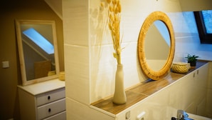 Ein Badezimmer mit einem Holzkosmetikschrank, einer Vase mit Trockenblumen, einem runden Spiegel und einer kleinen Pflanze auf der Ablagefläche.