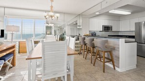 A modern kitchen with white cabinetry, a central island with bar stools, and a dining area with a wooden table and white chairs. - Ashworth 1801 4 Bedroom Condo (North Myrtle Beach)