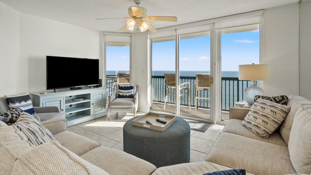 A spacious living room with a large flat-screen TV, comfortable seating, a ceiling fan, and a balcony with a view of the ocean. - Ashworth 1801 4 Bedroom Condo (North Myrtle Beach)
