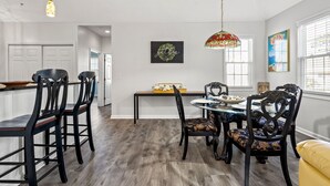 Un coin repas avec une table ronde, des chaises noires et un banc. On y trouve deux lampes suspendues, une guirlande accrochée au mur et une fenêtre avec des stores.