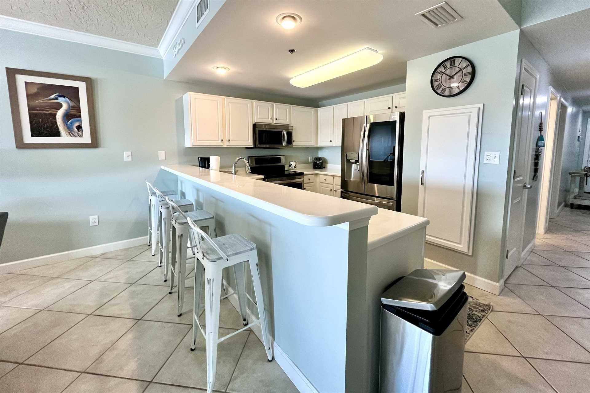 A modern kitchen with white cabinetry, a central island with bar stools, stainless steel appliances, and a clock on the wall.