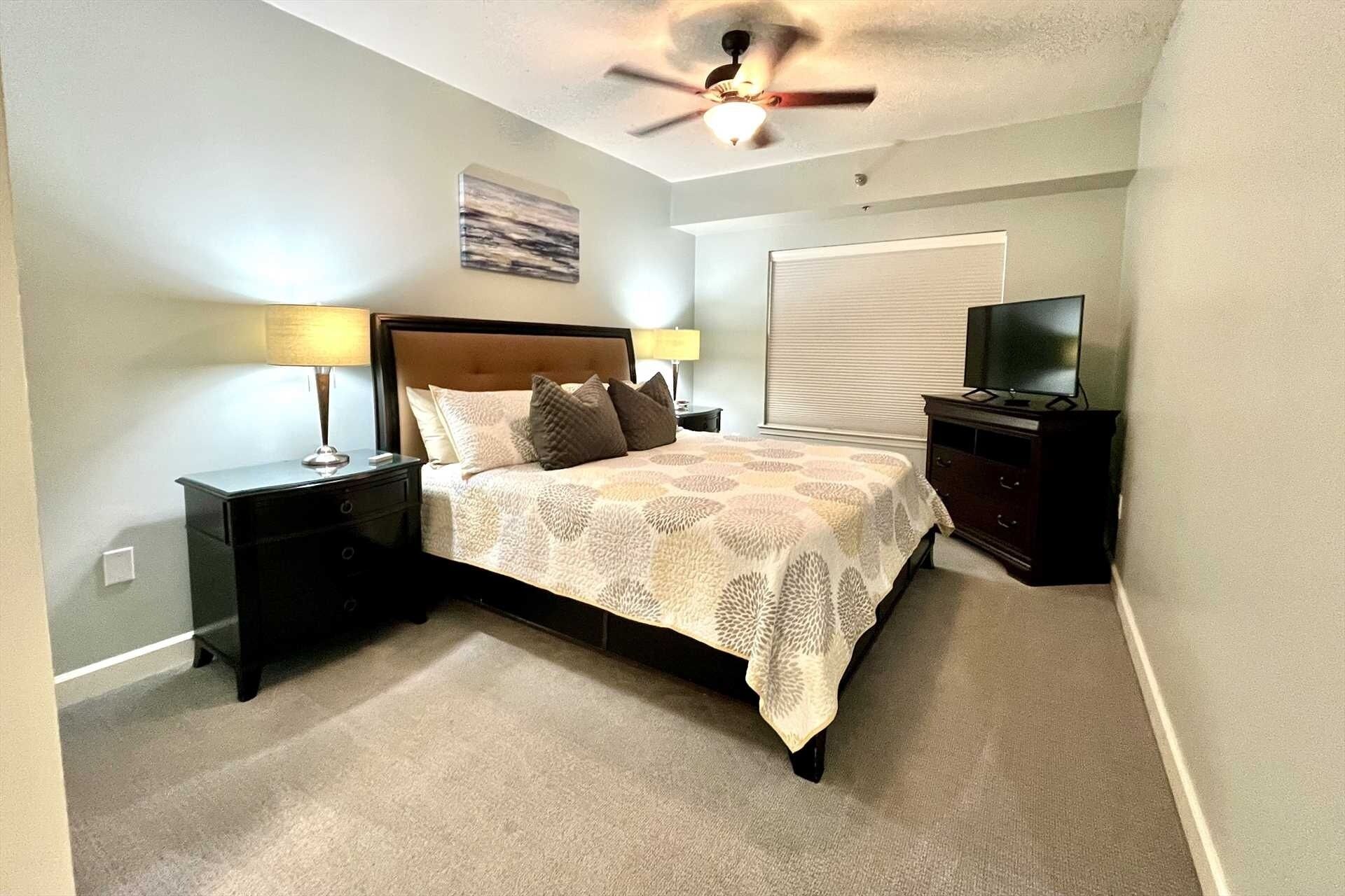 A bedroom with a bed, nightstands, a ceiling fan, a television, and a window with blinds.
