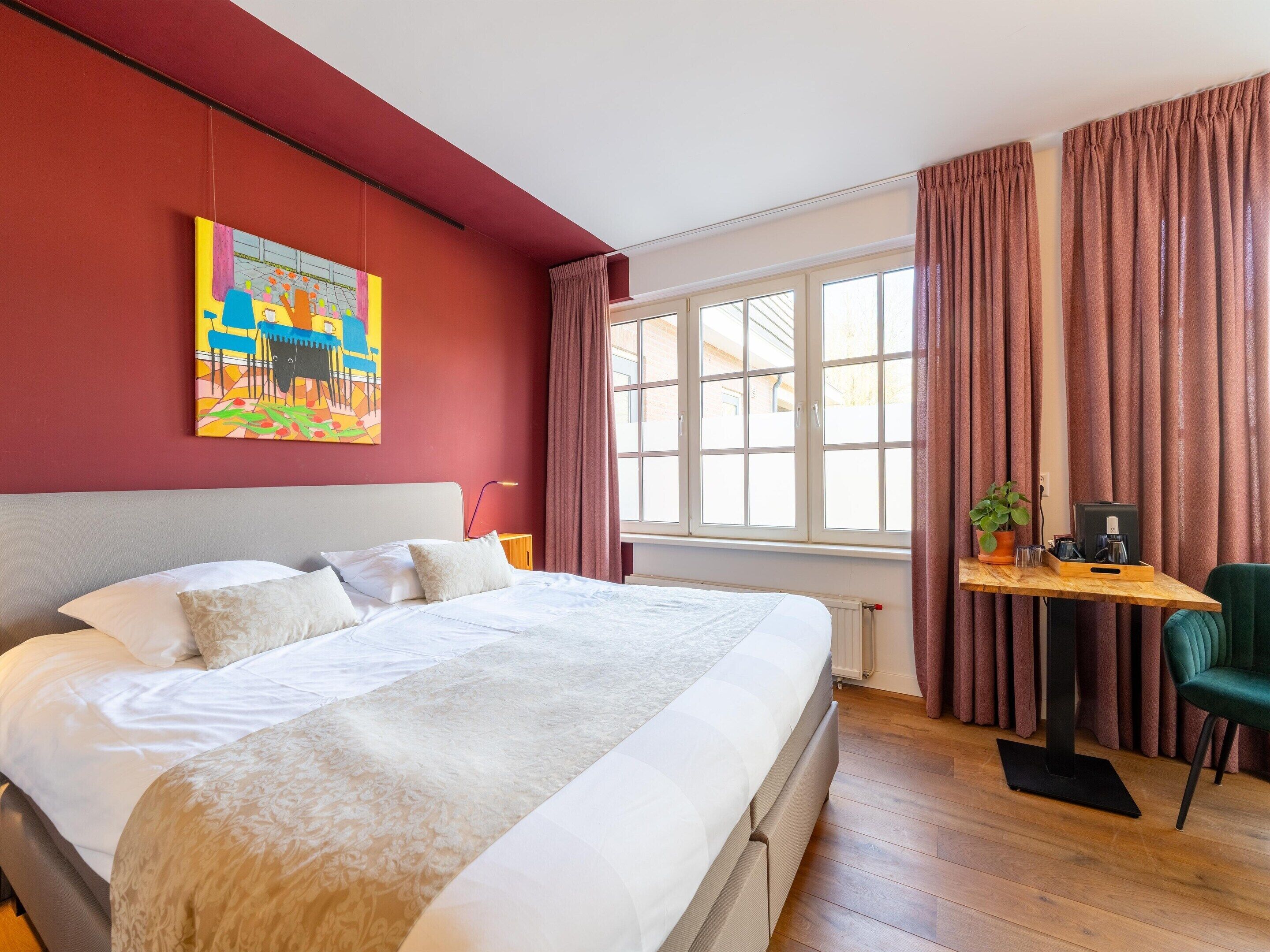 Quarto de hotel com uma cama grande, uma escrivaninha com computador, uma cadeira e uma janela com cortinas.