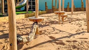 Hiekkainen leikkialue, jossa on puisia rakenteita, kuten liukumäki ja keinut, ja taustalla näkyy sinisellä maalattu rakennus.