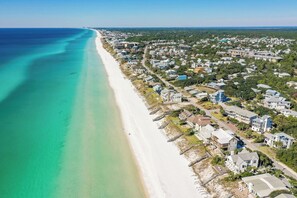 Vue aérienne d’une zone résidentielle côtière, avec des maisons le long d’une plage, une eau d’un bleu limpide et un ciel dégagé.