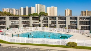 Ein modernes Gebäude mit einem Swimmingpool, umgeben von weißen Zäunen, mit Balkonen und freiem Blick auf hohe Gebäude im Hintergrund.