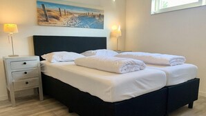 A neatly made bed with white linens and pillows, a bedside table with a lamp, and a painting of a beach scene on the wall. - Bungalow in Sint Maartenszee With Veranda (Sint Maartensvlotbrug)