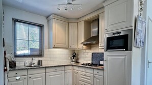 Une cuisine avec des meubles blancs, un four à micro-ondes, une cuisinière et une fenêtre avec des stores.