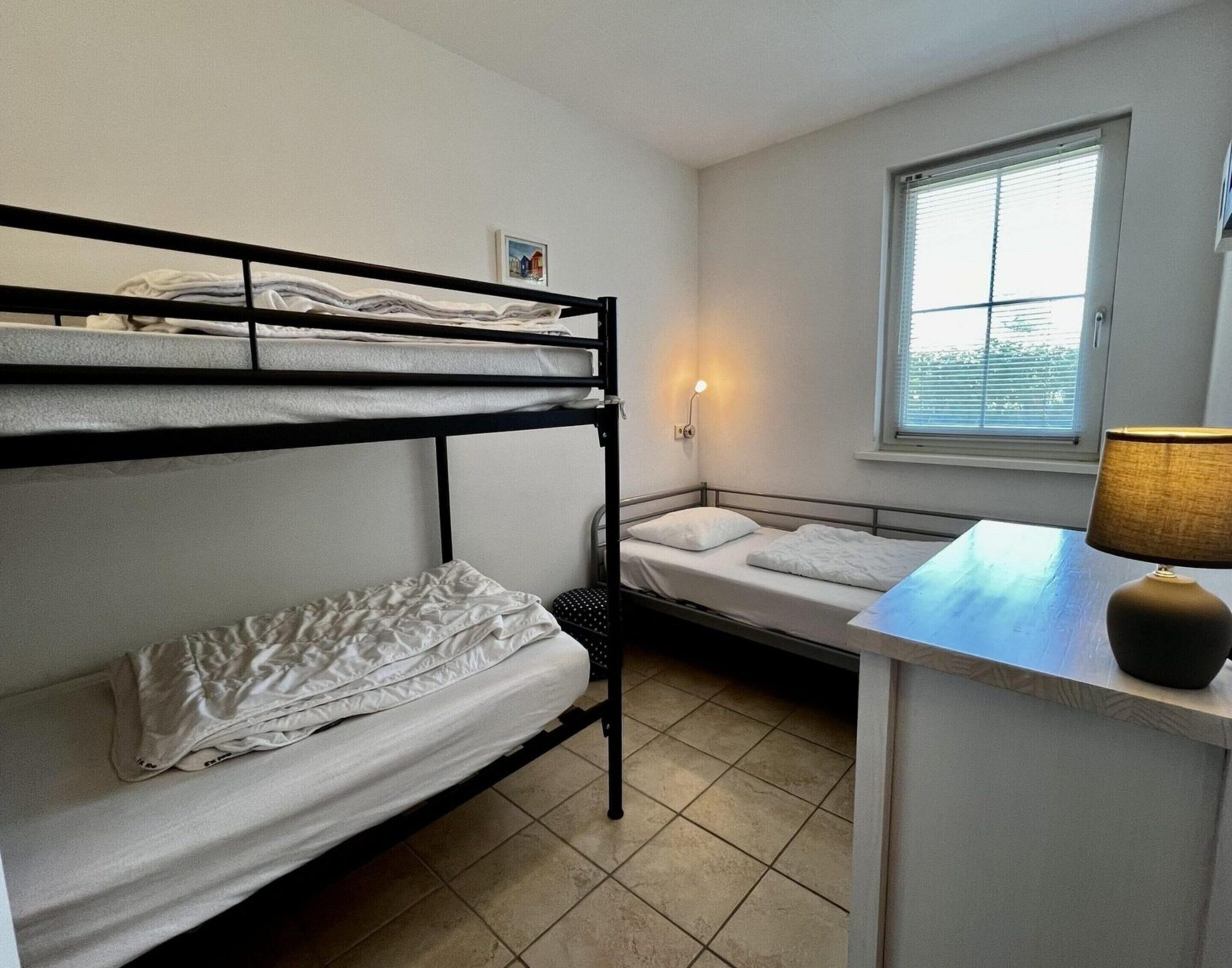 A dormitory room with bunk beds, a desk, a lamp, and a window with blinds.