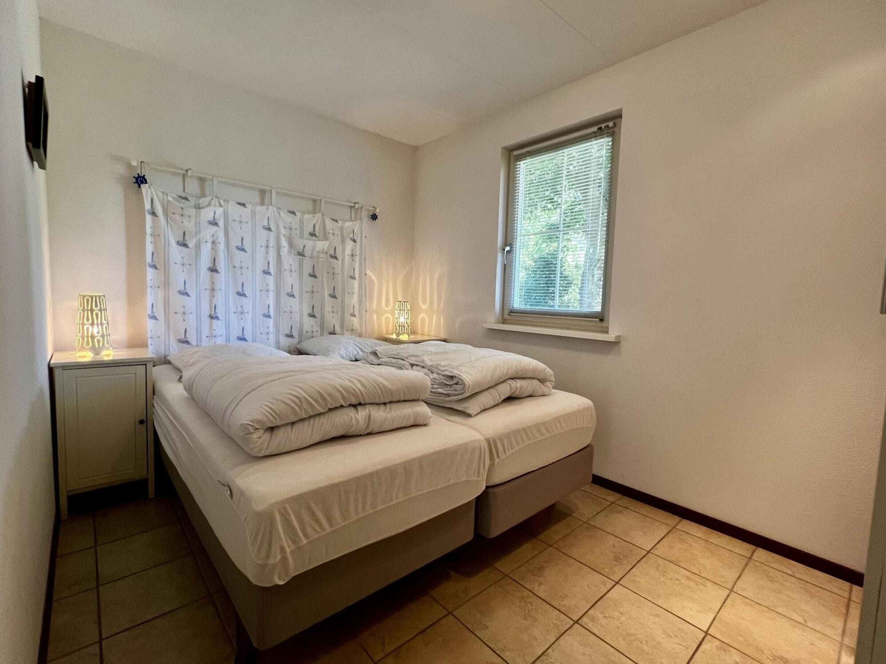 A bedroom with a bed, a nightstand, a window with blinds, and a wall-mounted light fixture.