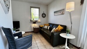 Un salon comprenant un canapé en cuir noir, un fauteuil en cuir noir, une petite table blanche, un lampadaire, un téléviseur, une fenêtre et une horloge murale.