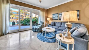 A spacious living room with a grey sofa, striped armchairs, a coffee table, and a floor lamp. A large window offers a view of a pool and a building with balconies. - Adagio D-103 Poolside 3/3 - NEW to program for 2025! (Santa Rosa Beach)