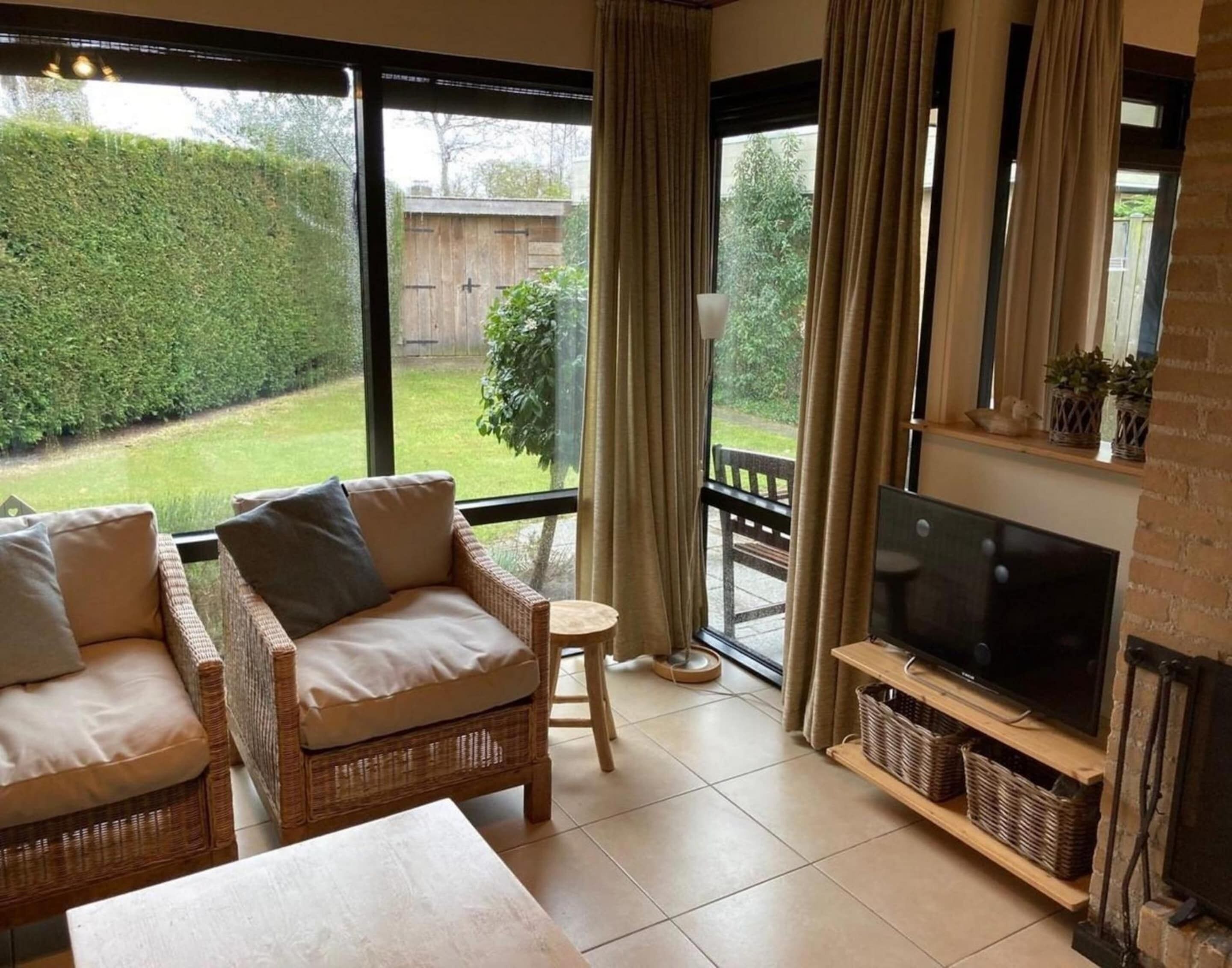 A cozy living room with wicker furniture, a flat-screen TV, and a view of a garden through large windows.