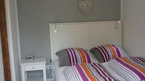 A bedroom with a white bed, striped pillows, a bedside table, a wall-mounted light, and a heart-shaped decoration. - Cosy Holiday Home Near Beach (Sint Maartensvlotbrug)