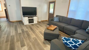 Ein geräumiges Wohnzimmer mit einem grauen Ecksofa, einem Flachbildfernseher auf einem weißen Standfuß, einem Deckenventilator und einem Holzboden.