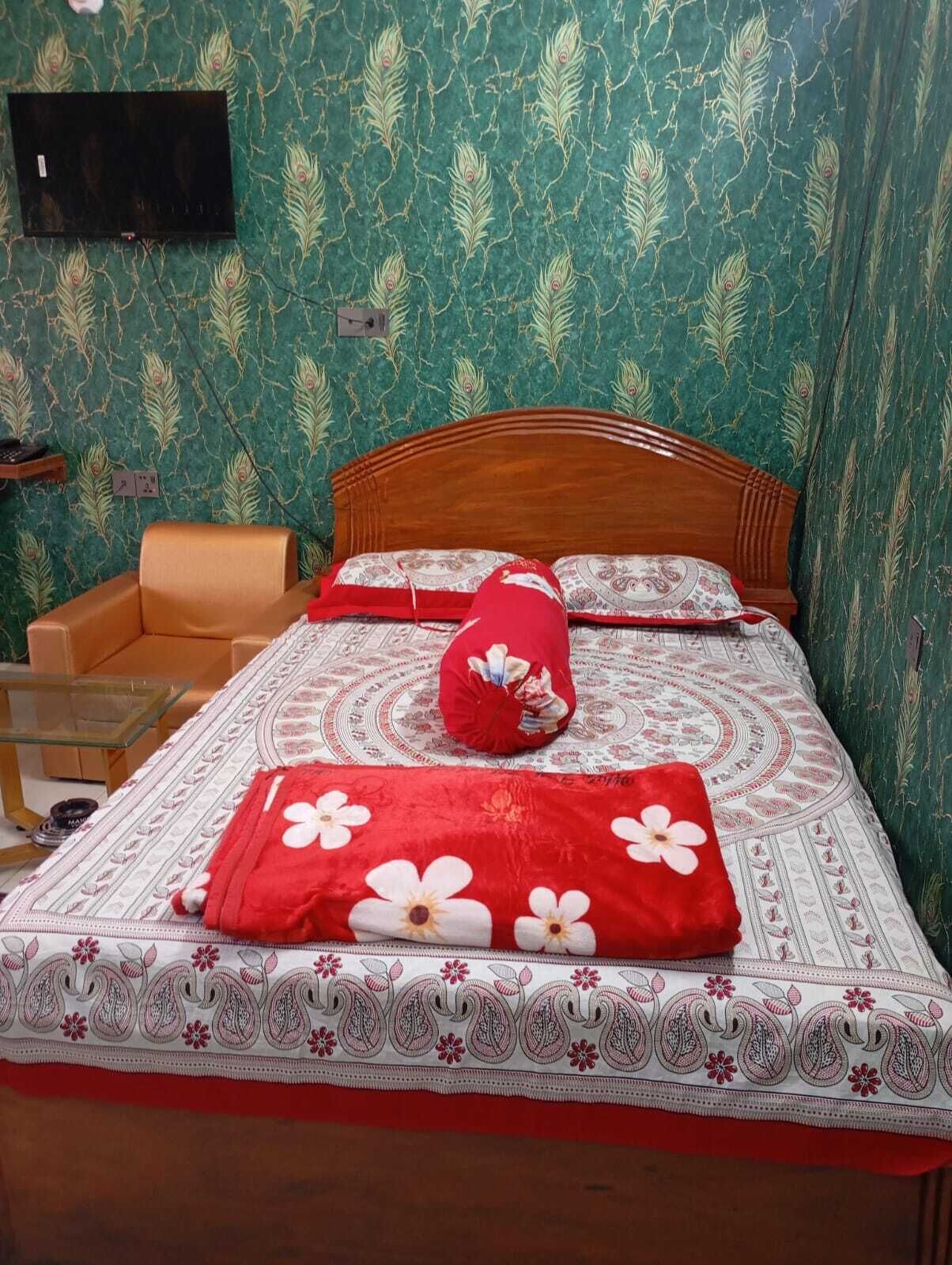 Siististi pedattu sänky, jossa on kukkakuviollinen päiväpeite ja tyynyjä, puinen pääty sekä vihreä tapetti, jonka kuvioinnissa on riikinkukon sulkia.