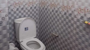 A bathroom with a white toilet, a red bucket, and tiled walls with a checkered pattern. - Hotel Moon International  (Dhaka)