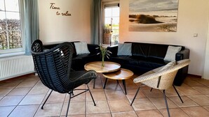 A living room with a black sofa, two wicker chairs, a small round table with a vase, and a painting on the wall. - Villa Close to Forest, Beach & sea (Sint Maartensvlotbrug)
