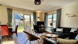 A spacious living room with a sofa, a dining area with chairs, a TV, and a view of the outdoors. - Villa Close to Forest, Beach & sea (Sint Maartensvlotbrug)
