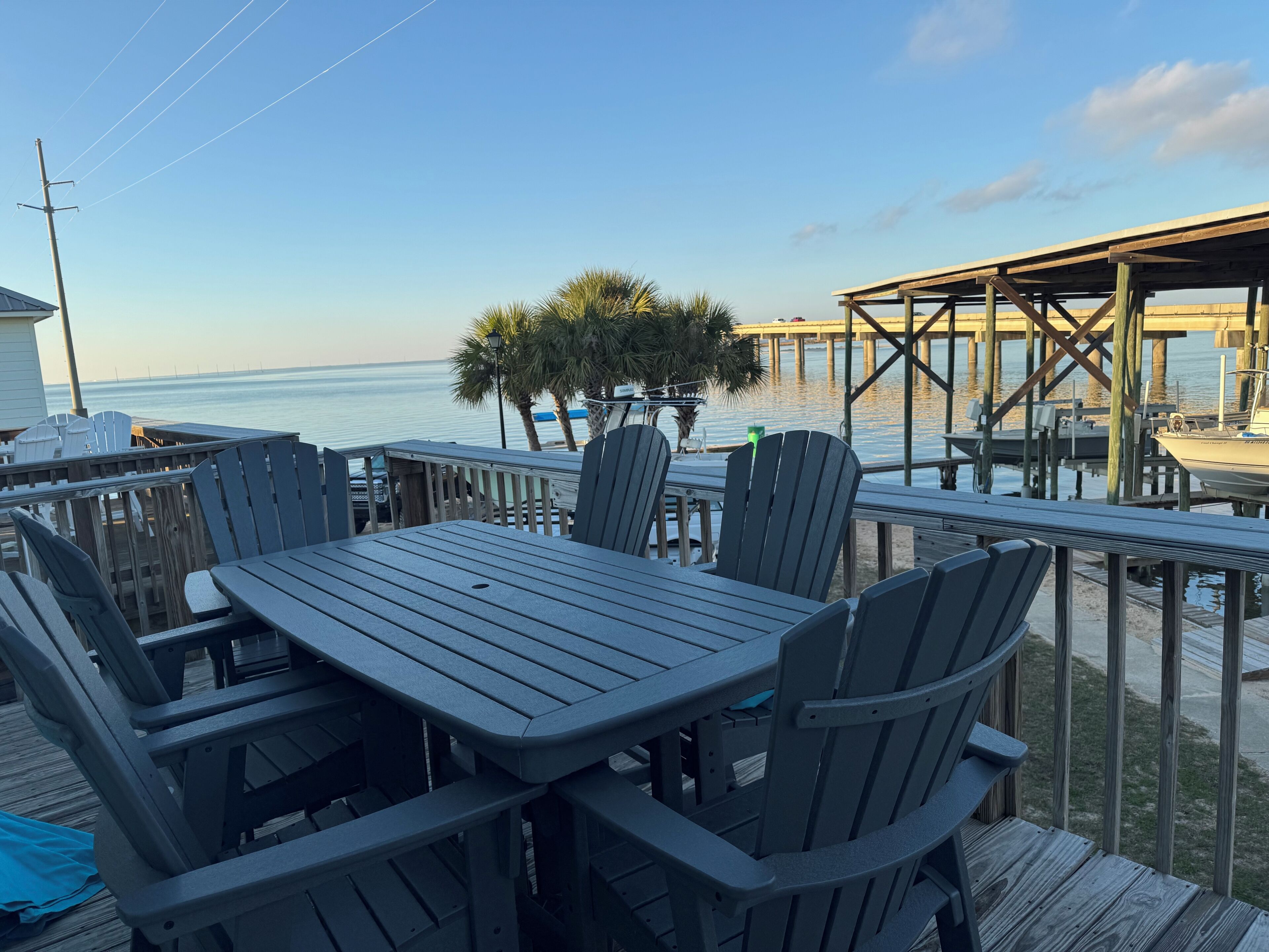 A wooden deck with a table and chairs, overlooking a calm body of water and a dock.