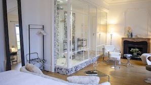 A modern hotel room with a large bed, a sitting area with a chair and a small table, a fireplace, and a large mirror.