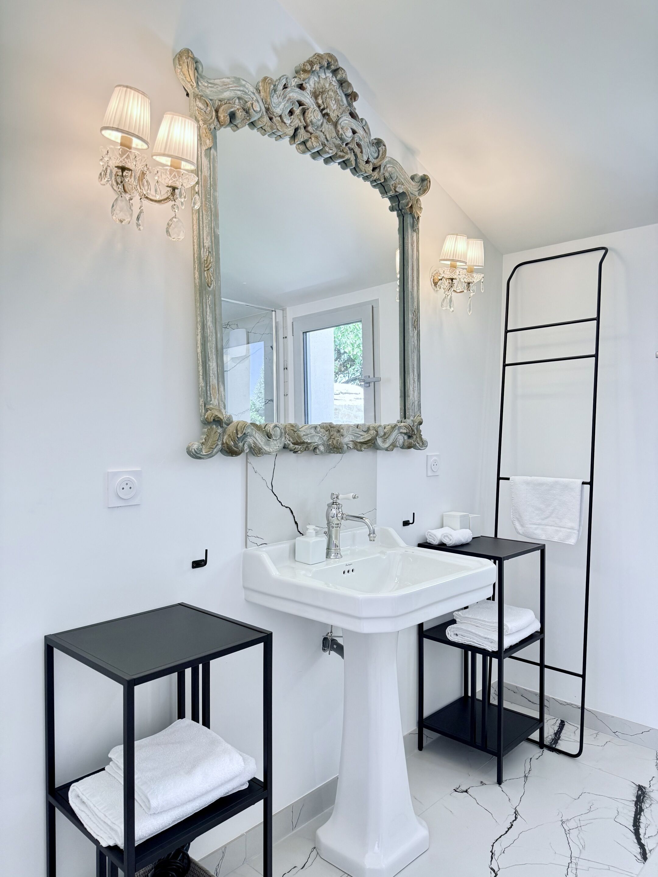 Een badkamer met een wit wastafeltje op voet, een grote, versierde spiegel, twee zwarte nachtkastjes met handdoeken, een handdoekhouder en een zwart metalen handdoekrek.