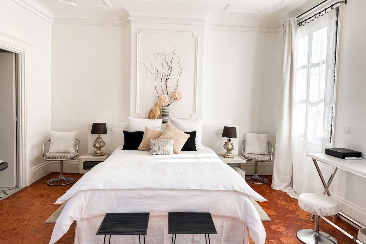 Habitación de hotel con una cama grande, dos mesitas de noche con lámparas, una silla, un taburete pequeño y una ventana con cortinas.
