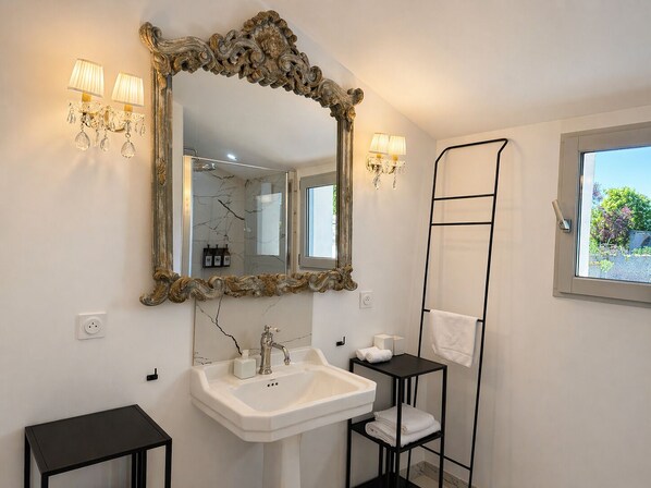 A bathroom with a large ornate mirror, a white sink, a black shelf with towels, and a window with a view.
