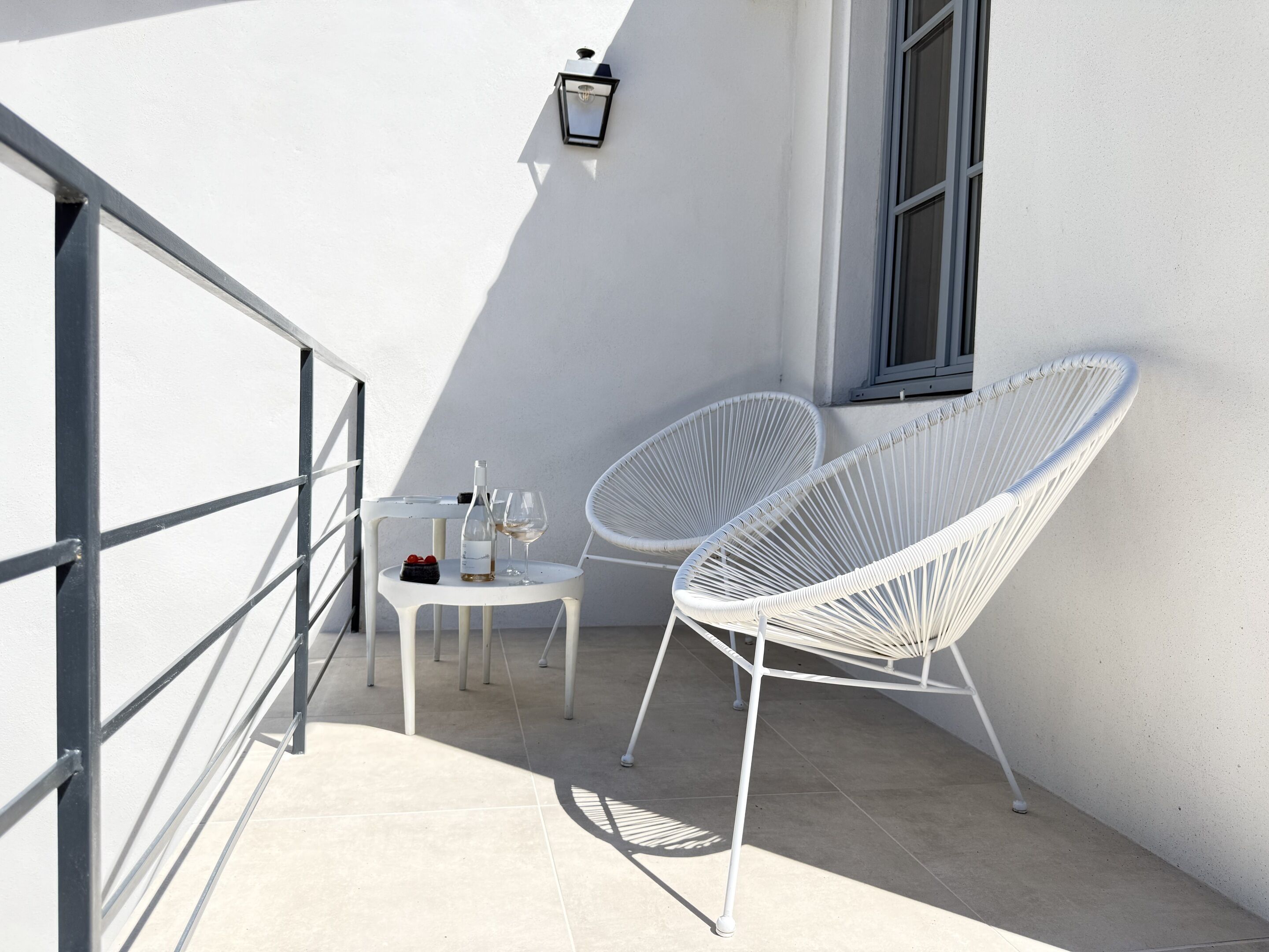 A balcony with white wicker chairs, a small table with a bottle and glasses, and a wall-mounted light fixture.