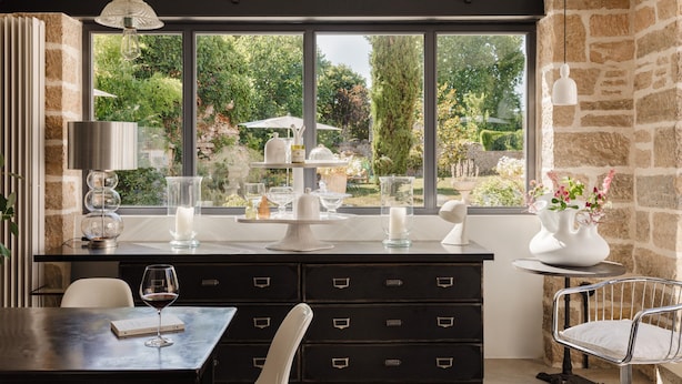 A modern dining area with a view of a garden through large windows, a black dining table with a wine glass, and a white chair.