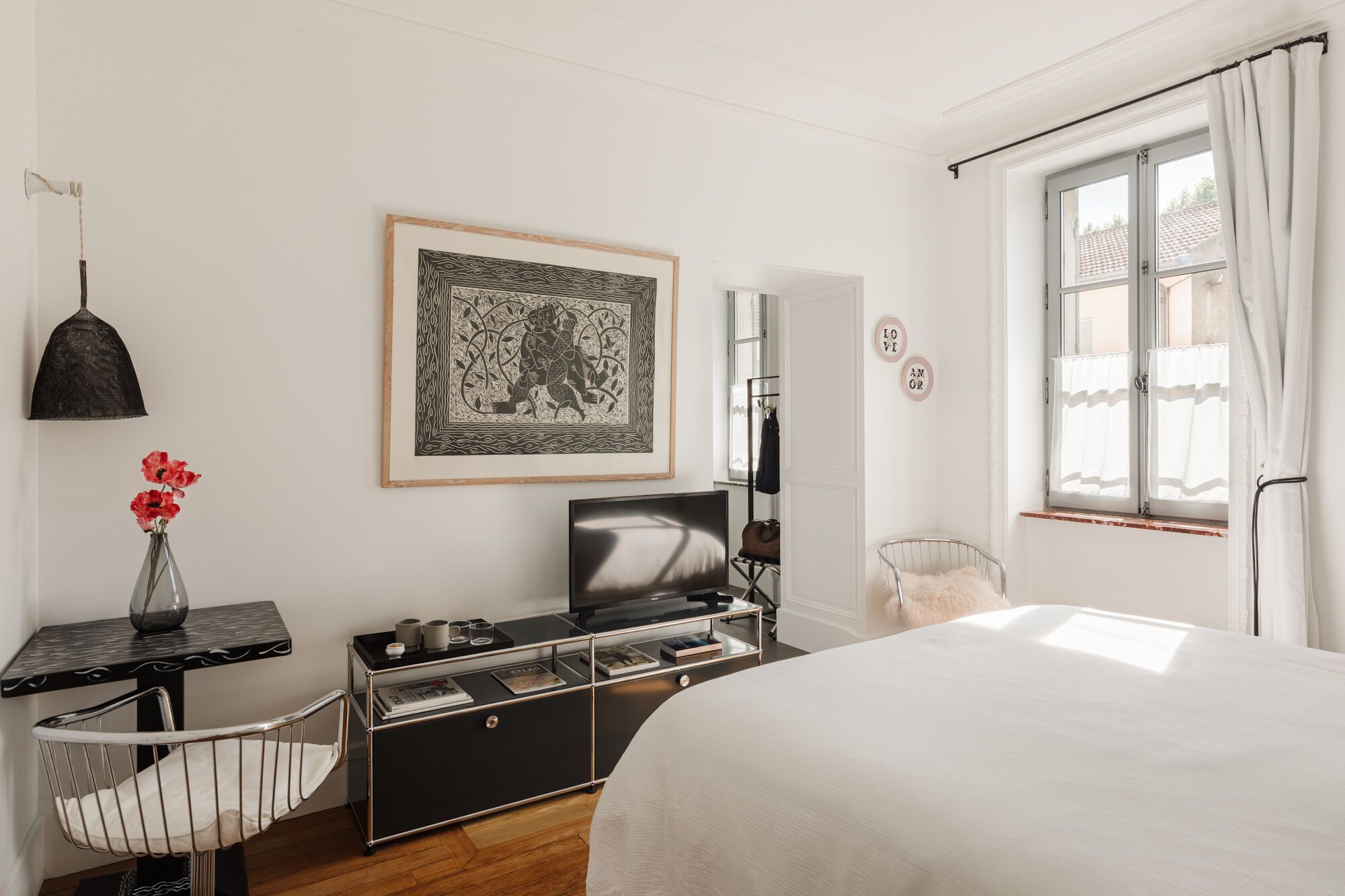 Een slaapkamer met een bed, een bureau met stoel, een televisie, een lijst met kunstwerk, een hanglamp en een raam met gordijnen.
