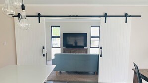 A modern kitchen with a white countertop, black stools, and a sliding barn door leading to a living area with a TV. - Modern & Spacious 2yo Home (New Plymouth)