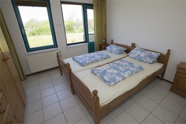 A double bed with floral bedding, a window with a view of greenery, and a radiator under the window. - Furnished Apartment on the Dunes (Sint Maartensvlotbrug)