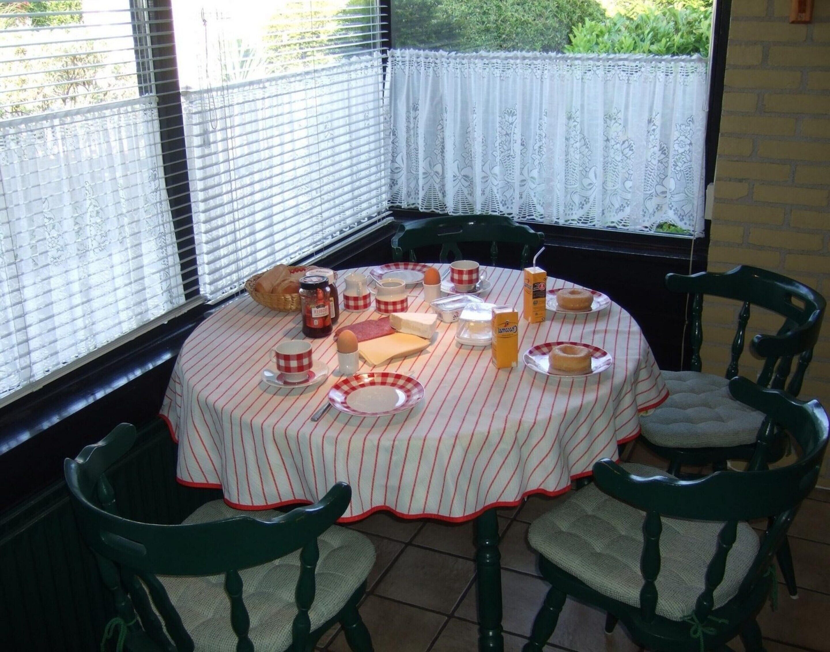 Une table ronde recouverte d’une nappe à rayures, entourée de quatre chaises. La table est mise avec des assiettes, des tasses, une bouteille et un panier de pain.