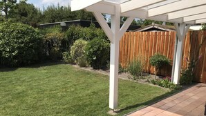 Een achtertuin met een houten omheining, een witte pergola en een hond die op het terras zit.