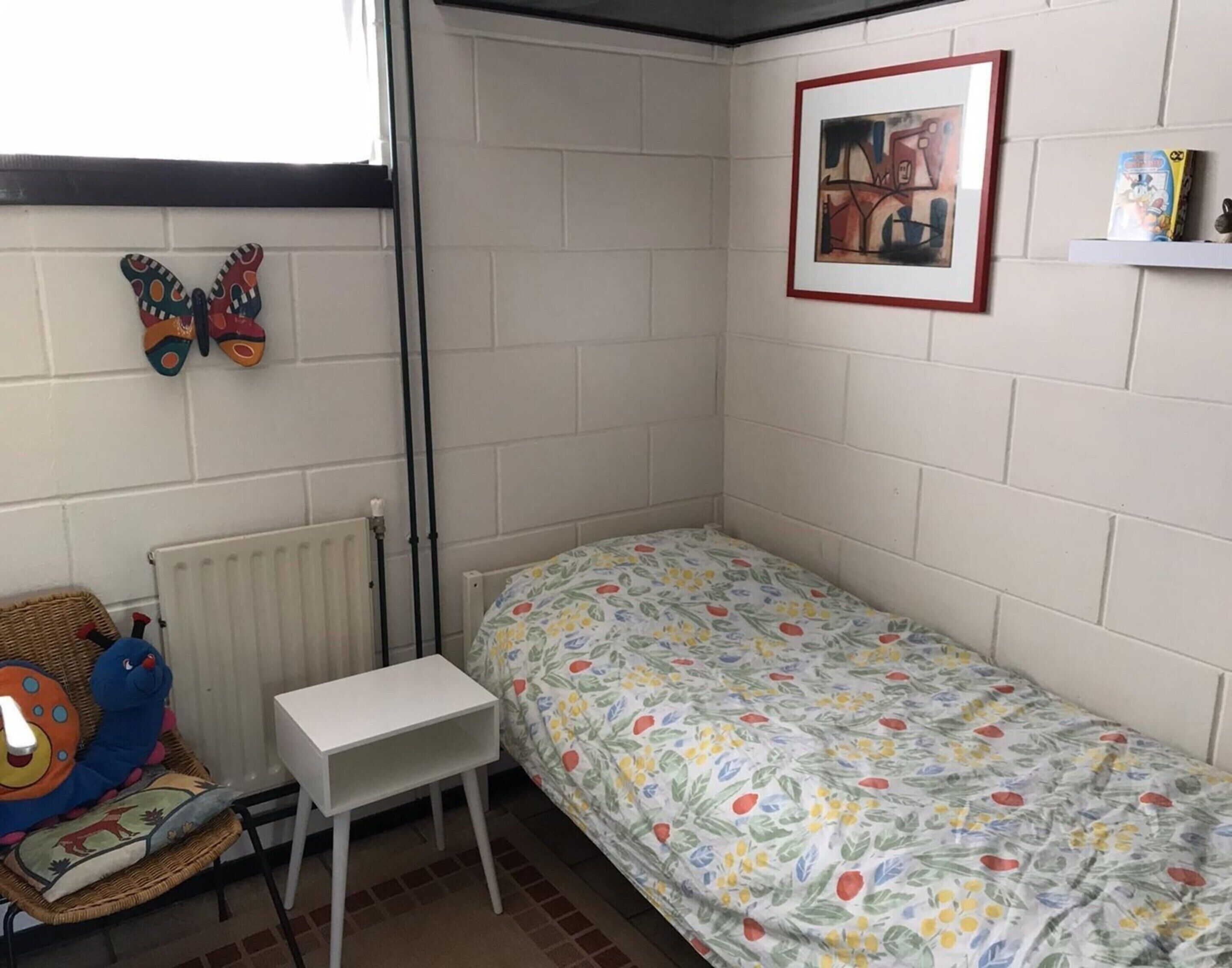 A small, single-bed room with a white brick wall, a radiator, a small white table, a wicker chair, and a colorful bedspread.