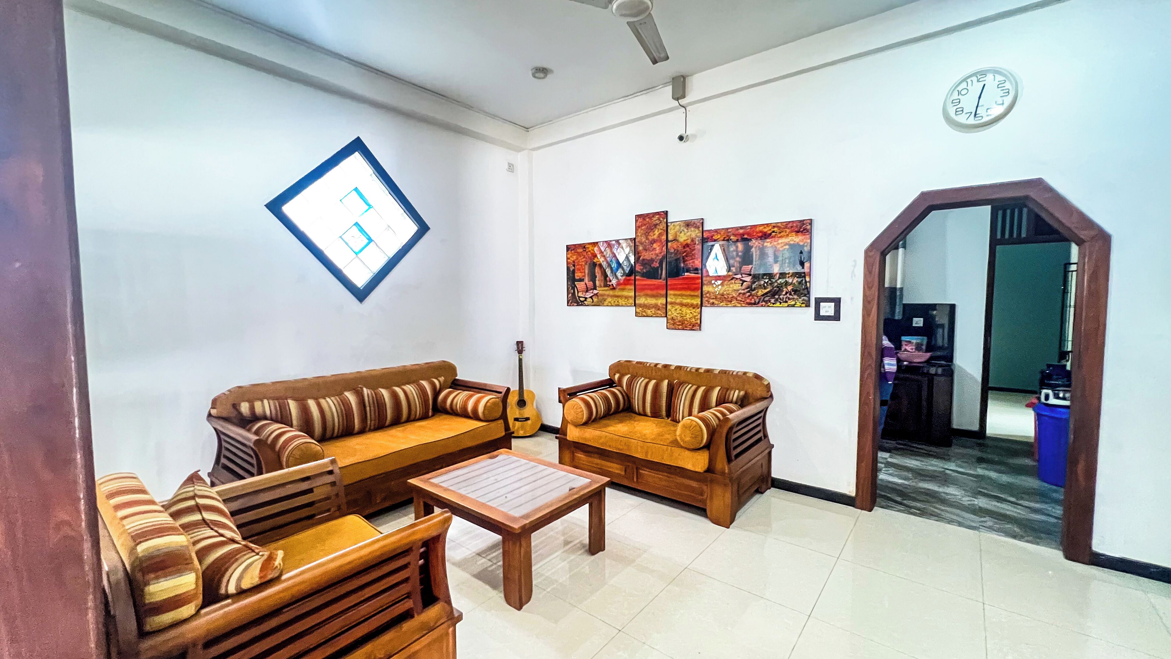 Una sala de estar con dos sofás, una mesa de centro y una guitarra. Se observan pinturas colgadas en la pared y un reloj.