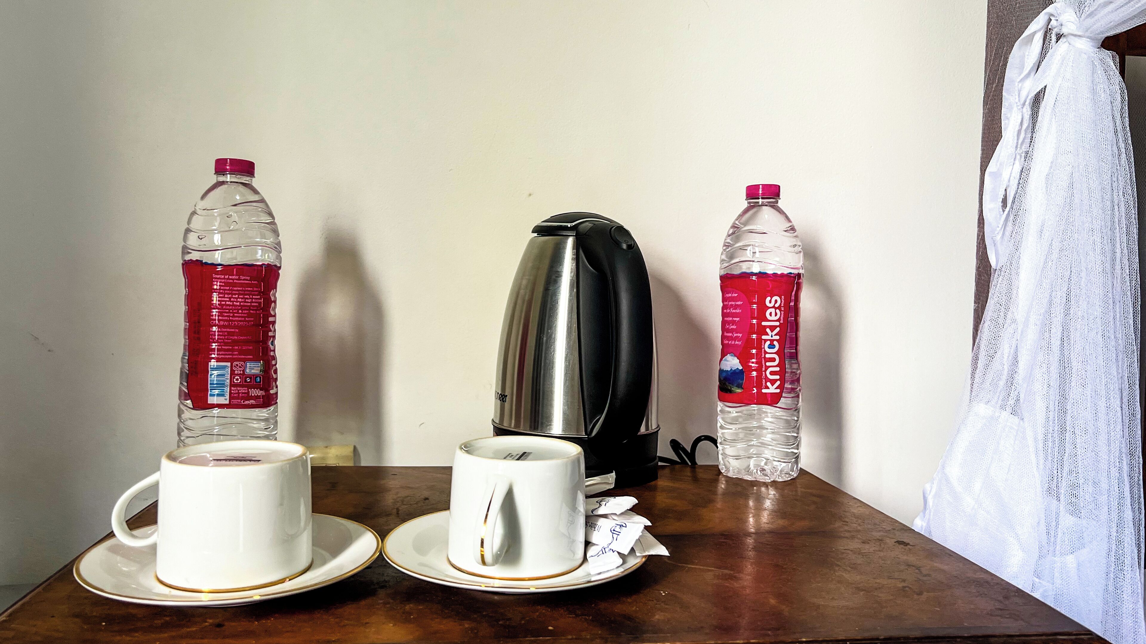 Una mesa con dos tazas blancas en platillos, una tetera eléctrica de acero inoxidable y dos botellas de agua con etiquetas rojas.