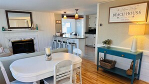 A dining area with a white table and chairs, a blue console table with baskets, a fireplace, and a kitchen in the background. - Easy Walk to West Dennis Beach, AC, Linens, Kayaks & SUP (Dennis)