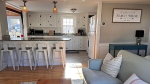 Une cuisine moderne avec des armoires blanches, un îlot central doté de tabourets de bar, un coin repas avec un canapé et une table basse, et une affiche sur le mur indiquant : « MAISON DE PLAGE, fondée en 2024 ».