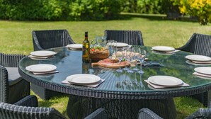 Een ronde glazen tafel voor zes personen, voorzien van rieten stoelen, een fles, een karaf en een schaal met fruit.