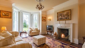 Een traditionele woonkamer met een open haard, comfortabele zitplaatsen en een groot raam.