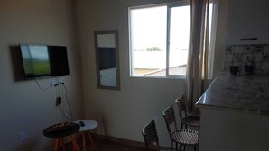 Una habitación pequeña y bien iluminada, con un televisor montado en la pared, una mesa redonda pequeña con una silla, y una ventana que ofrece vistas al exterior.