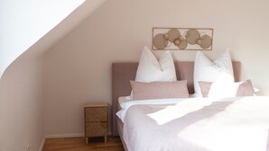 Un dormitorio con techo inclinado, una cama con varias almohadas, una mesita de noche pequeña y una obra de arte colgada en la pared.