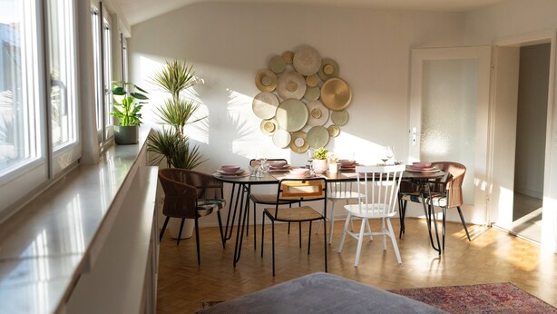 Un comedor con una mesa lista para cuatro personas, rodeada de sillas, un arreglo decorativo de platos en la pared y plantas en macetas.