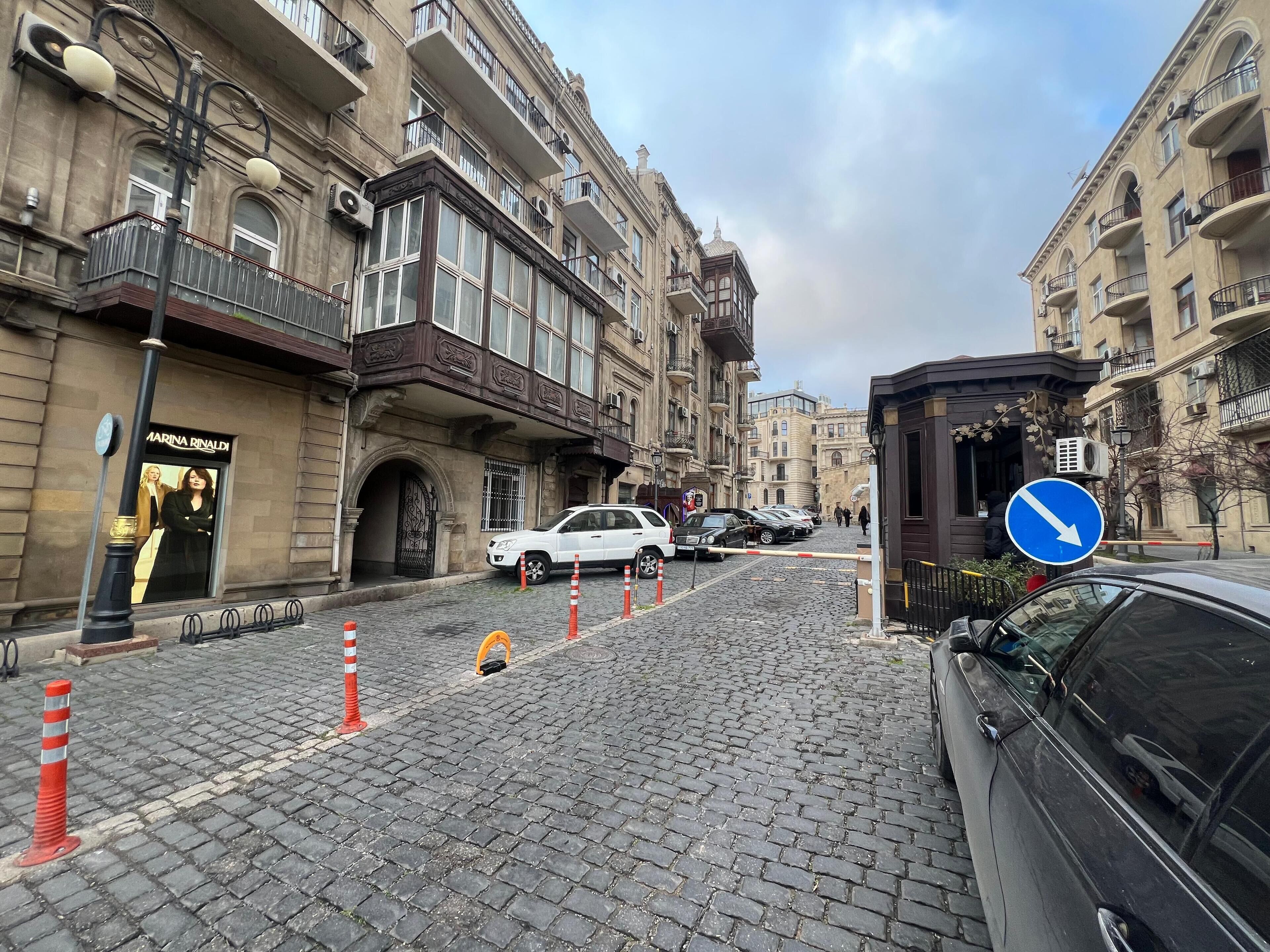 A cobblestone street with historic buildings, a white car parked on the side, and a blue directional sign.