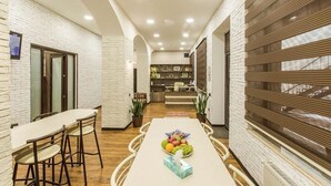 A modern dining area with a long table, chairs, and a fruit bowl. There is a kitchen area in the background with a counter and appliances. - Salam Old Baku Hostel (Baku)