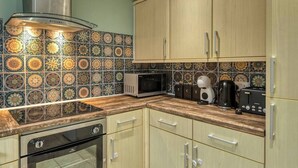 A kitchen with a microwave, toaster, and coffee maker on the counter, with patterned tiles on the backsplash and wooden cabinets. - Cosy 2BDR Trafford Cottage, Fits 5, EV charger, WIFI, Parking 
 (Partington)